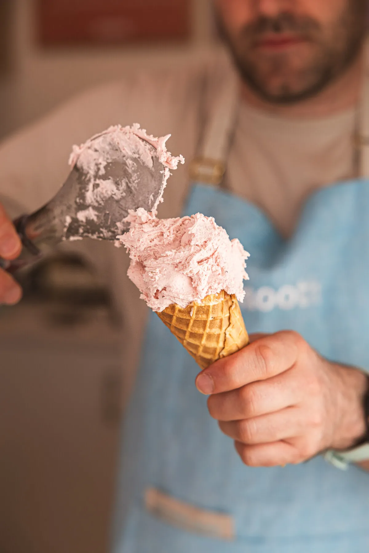 Hand-scooping strawberry gelato onto a waffle cone, Smooth apron in the background.
